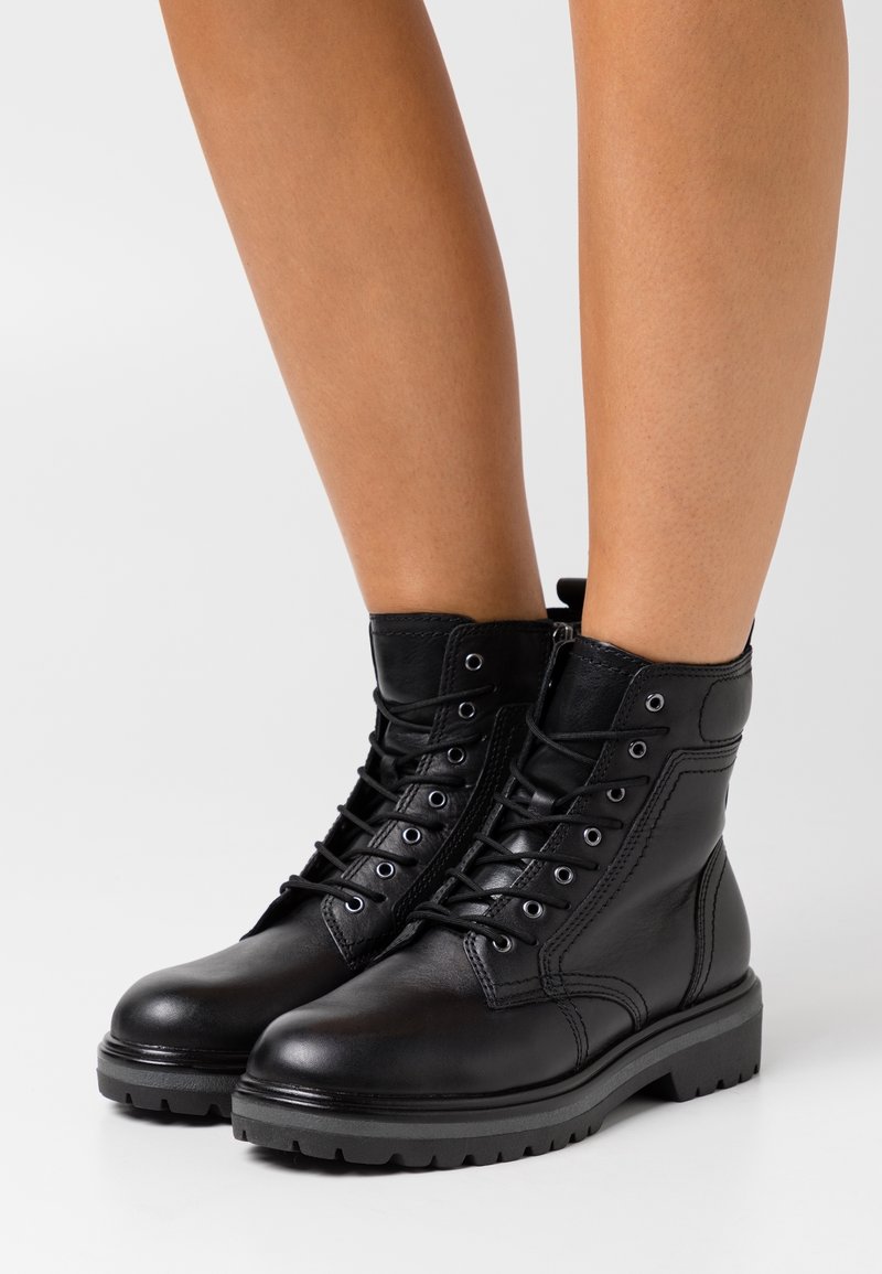Botines de cuero negro hasta el tobillo con cordones en la parte delantera, puntera redonda y suela de goma texturada. Presenta ojetes metálicos y un diseño cosido.