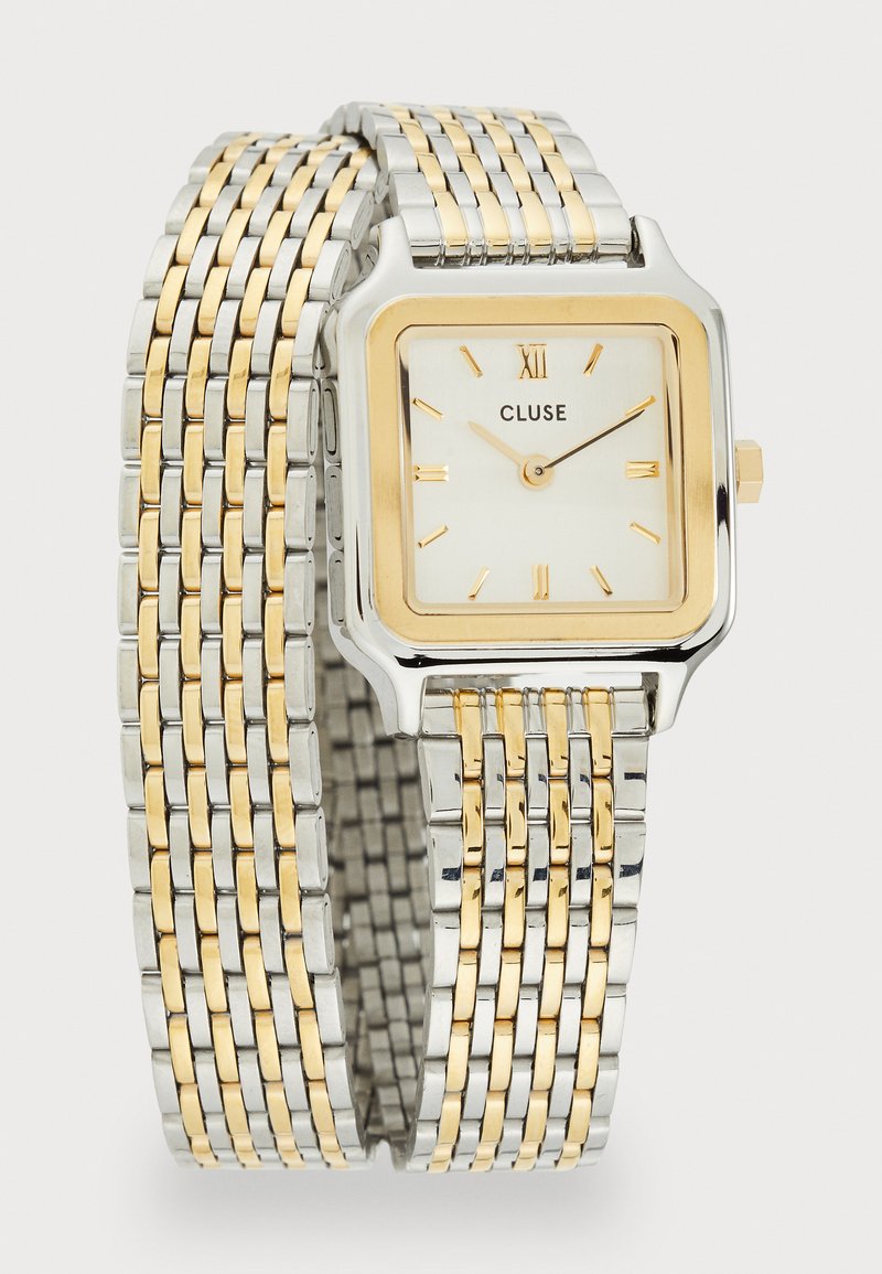 Ruudu kuju hõbe- ja kuldtooni käekell valge näo, kuldsete tundide markerite ning vahelduvate hõbedaste ja kuldsete linkidega kudumetallist rihmaga.