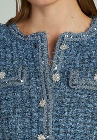 Giacca blu con texture e motivo in tweed, caratterizzata da accenti in paillettes e bottoni decorativi sul davanti e sul colletto.