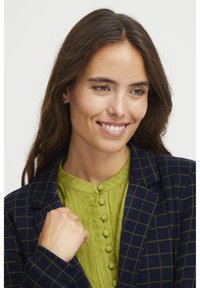 Tumed blazer rohelise ruudustikuga, mille all on tekstuuriga roheline pluus, millel on ümar kaelus ja nööbiekraanid. Looduslikud juuksed ja hele nahatoon.