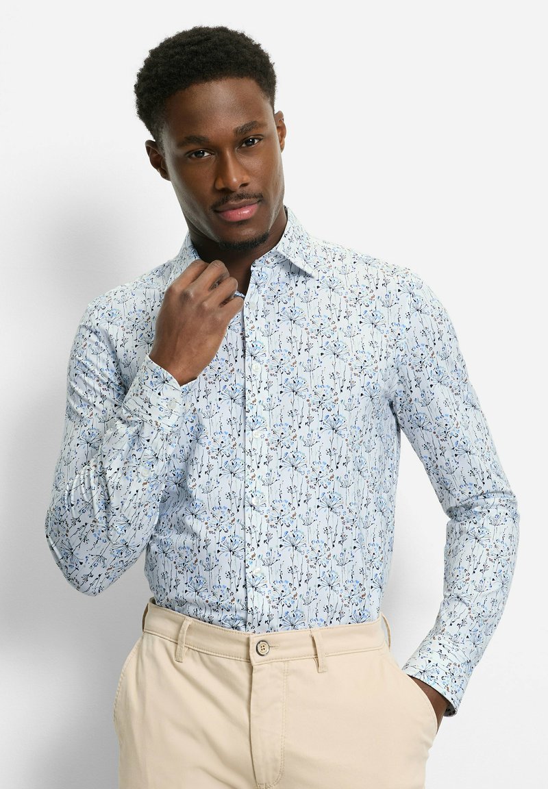 Mann, der ein hellblaues, floral gemustertes Hemd mit Knöpfen und beige Hosen trägt, posiert mit einer Hand, die den Kragen richtet, und der anderen in der Hosentasche.