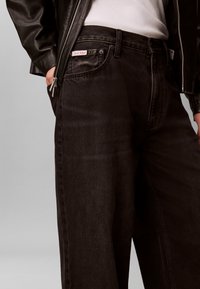 Άτομο που φοράει μαύρα τζιν Calvin Klein, λευκό πουκάμισο και μαύρο δερμάτινο μπουφάν, με το ένα χέρι στην τσέπη, σε γκρι φόντο.