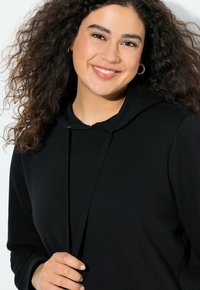 Donna sorridente con capelli ricci scuri che indossa orecchini a cerchio dorati e una felpa nera, che tiene i lacci della felpa contro uno sfondo chiaro.
