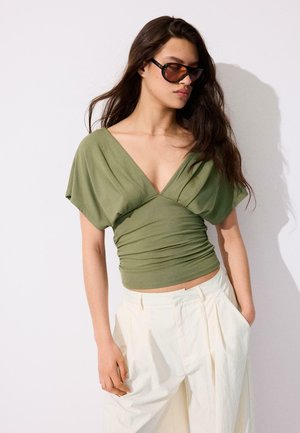 Žena s dlhými tmavými vlasmi nosiaca veľké hnedé slnečné okuliare, olivovo zelené tričko s hlbokým výstrihom do V a voľné biele nohavice s vysokým pásom.