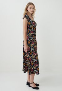 Blumen-Midi-Kleid mit einem navy-blauen Hintergrund, kurzen Ärmeln und einer taillierten Taille. Bunte Blumen bilden ein lebhaftes Muster. Es werden schwarze Ballerinas getragen.