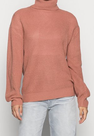 Personne portant un pull à col roulé large en tricot rose et un jean gris clair, debout devant un fond uni.