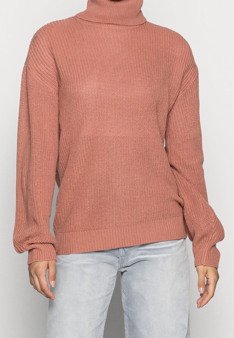 Personne portant un pull à col roulé large en tricot rose et un jean gris clair, debout devant un fond uni.