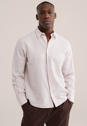 Man draagt een lichtroze overhemd met lange mouwen en kraag, donkerbruine broek, staand met handen in de zakken tegen een neutrale achtergrond.