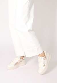 Weiße Sneakers mit floralen Stoffakzenten an den Seiten, strukturiertem Gummisohle und runder Zehenpartie. Die Schnürsenkel haben verstärkte Ösen für zusätzliche Haltbarkeit.