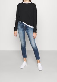 Černá mikina s dlouhým rukávem přes bílou košili, v kombinaci s modrými džínami středního vzestupu a bílými teniskami. Hladká textura, přiléhavý design.