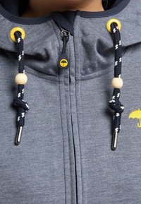 Sweat à capuche gris avec des cordons noir et blanc ornés de perles en bois, œillets jaunes et un petit logo d'un parapluie jaune près de la tirette.