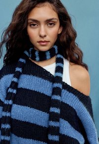 Blau-schwarz gestreifter Strickpullover mit einem schulterfreien Design, begleitet von einem gestreiften Schal in ähnlichen Farben und Textur.