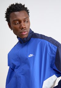 Zils Nike mētelis ar augstu apkakli, balta emblēma, kontrastējošas tumšas piedurknes un gluda tekstūra. Iekļauj rāvējslēdzēja aizdari un sportisku dizainu.