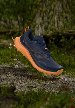Chaussure de trail bleu foncé et orange suspendue au-dessus d’un sol rocheux avec un fond de forêt verte.