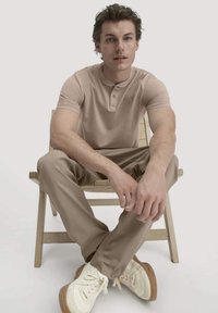 Beige Poloshirt mit Knopfleiste, kombiniert mit hellbeigen Hosen. Model sitzt auf einem Holzstuhl und trägt weiße Sneakers.