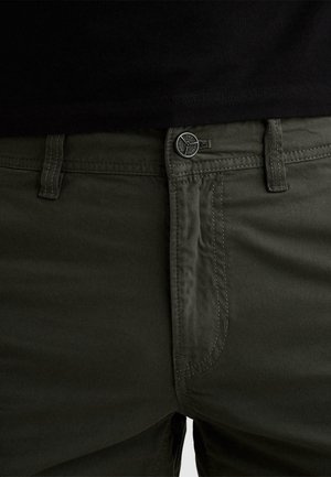 Tmavě zelené kalhoty s knoflíkovým zapínáním a poutky na pásek, nošené s černým topem částečně viditelným v pase.