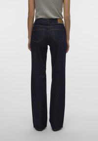 Jeans de mezclilla azul oscuro con corte recto, costuras visibles y dos bolsillos traseros; la cinturilla presenta una parche de cuero.