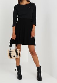 Schwarzes Langarmkleid mit taillierter Oberteil und schwingendem Rock, kombiniert mit einer karierten Tasche mit Fransenakzenten und schwarzen Ankle-Boots.