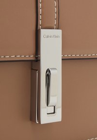 Kožená taška světle hnědé barvy s kovovou sponou, na které je vyryto "Calvin Klein". Okraje jsou zdobeny bílým prošíváním.