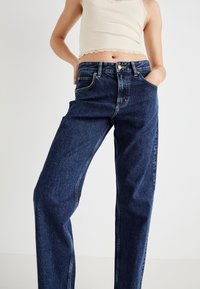 Oseba v temno modrih visokih kavbojskih hlačah in kremni brezrokavnici z čipkastim robom, z rokami v žepih kavbojk na enostavni ozadju.