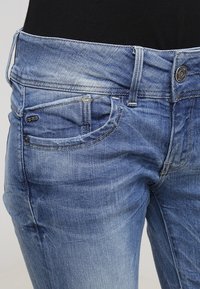 Jeans de mezclilla azul claro con un lavado desgastado, que presentan un diseño de cinco bolsillos, presillas para el cinturón y herrajes metálicos en la parte frontal.