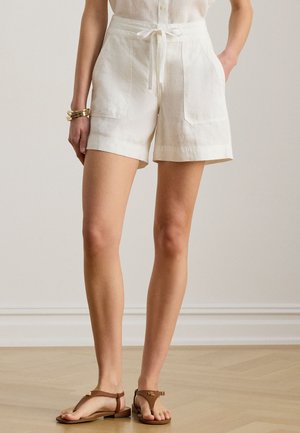 Žena nosiace biele ľanové šortky s vysokým pásom a prednými vreckami, s opaskom na zaväzovanie, béžovú košeľu, zlaté náramky a hnedé sandále na plochom podpätku.