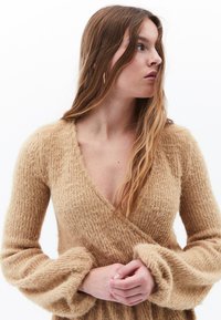 Beige strukturerad stickad tröja med djup V-ringning, långa puffy ärmar och mjukt, fluffigt material. Har en omslagsdesign för extra stil.