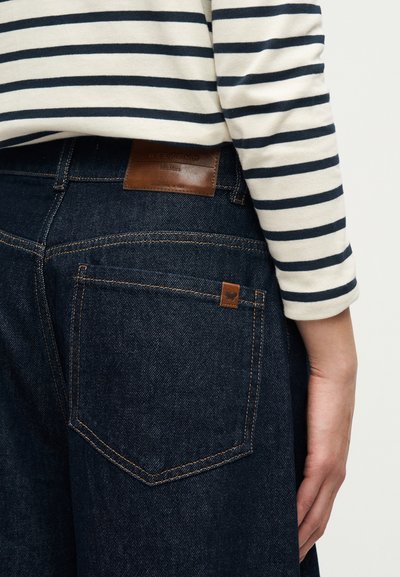 Άτομο που φοράει σκούρο μπλε τζιν με καφέ δερμάτινη ετικέτα και πίσω τσέπη, σε συνδυασμό με μακρυμάνικο λευκό πουκάμισο με μαύρες ρίγες.