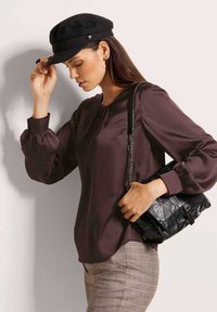 Frau passt schwarze Mütze an, trägt eine dunkelviolette Bluse mit Puffärmeln, karierte Hose und hält eine schwarze Leder-Schultertasche mit Kettenriemen. 