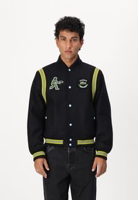 Veste universitaire noire avec des poignets rayés jaunes et bleus, présentant un joueur de tennis brodé en vert et le logo Lacoste. Texture de tissu douce.