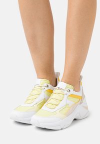 Weiße und hellgelbe Sportschuhe aus Mesh- und synthetischem Material, mit rundem Zehenbereich, markanter Sohle und Schnürsystem mit gelben Akzenten.