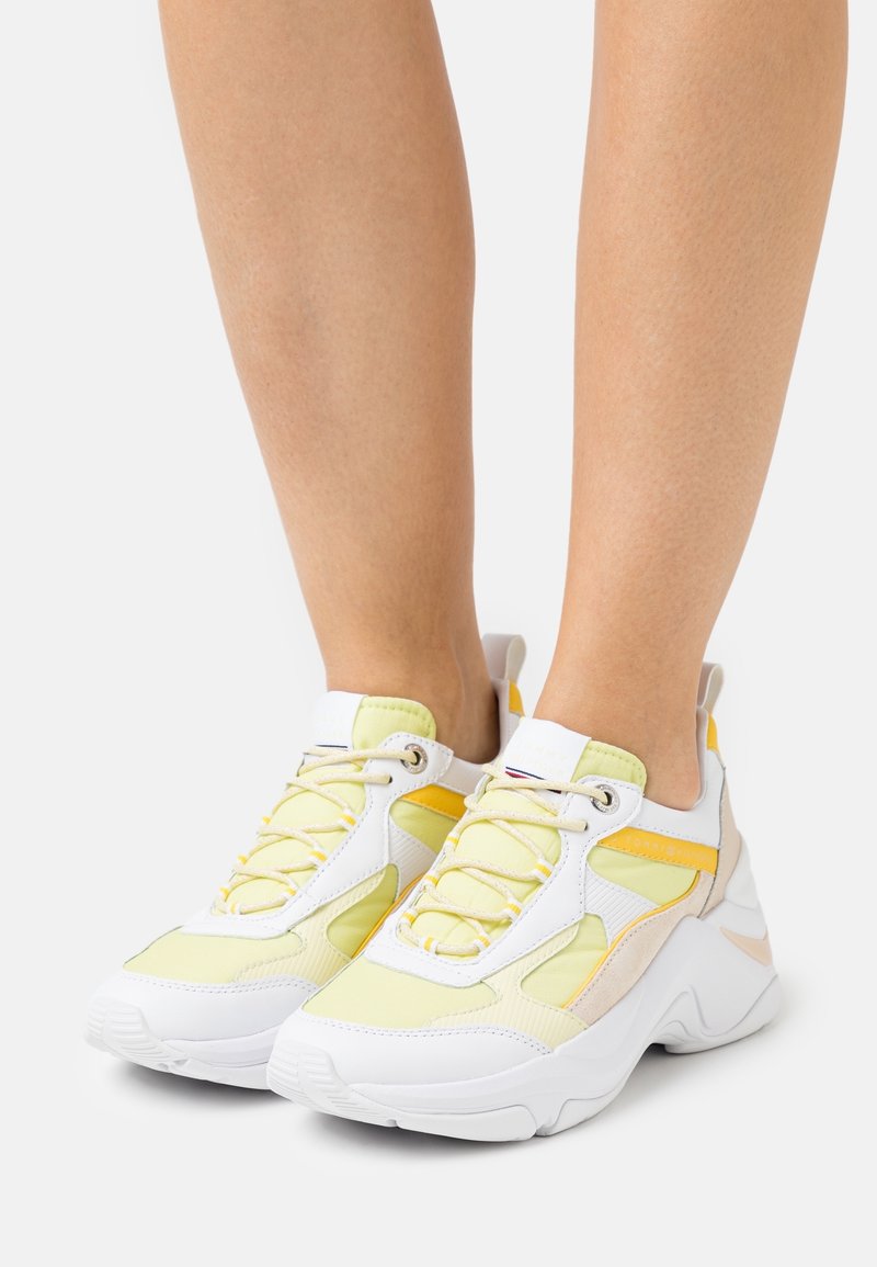 Weiße und hellgelbe Sportschuhe aus Mesh- und synthetischem Material, mit rundem Zehenbereich, markanter Sohle und Schnürsystem mit gelben Akzenten.