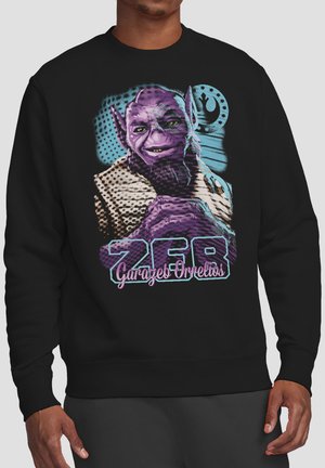 Mann trägt schwarzen Sweatshirt mit farbigem Grafikdruck eines violetten Alien-Charakters und dem Text "Zer Garazeb Ornellios" auf der Vorderseite.