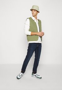 Colete acolchoado verde sobre uma camisa de manga longa branca, jeans azul-escuro e ténis verdes e brancos. Inclui um chapéu de balde de cor clara.