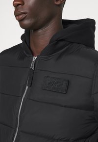 Schwarze Pufferjacke mit Kapuze, mit Reißverschluss, strukturierten Nähten und einem Patch-Logo auf der Brust. Glatte Stoffoberfläche.