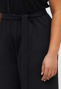 Black textured fabric pants with a tied waist belt. Features a loose fit and subtle patterned surface, showcasing a smooth hand feel.
