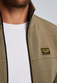 Olijfgroene zip-up jas met een gestructureerd oppervlak, zwarte rits en een gouden logo op de linkerborst. Witte t-shirt eronder.