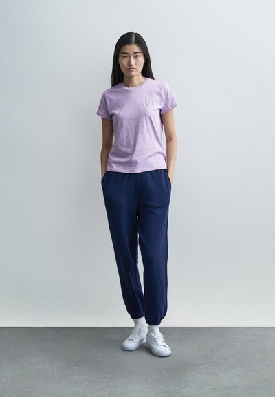 Lilla t-skjorte med korte ermer kombinert med marineblå joggebukser. Antrekket har en avslappet passform og hvite joggesko. Jevn stofftekstur.