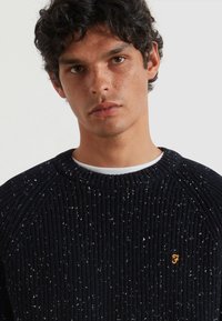 Joven con cabello corto y rizado de color oscuro que lleva un suéter de punto negro texturizado con pequeñas motas blancas y un logotipo naranja de "F" en el pecho.
