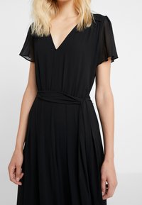 Vestido negro con escote en V, mangas cortas abullonadas y un cinturón de tela. Está hecho de un material ligero y cuenta con una falda fluida con pliegues.