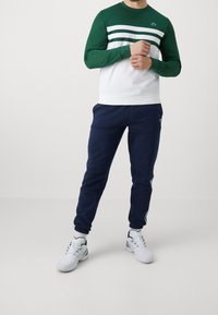 Grön och vit långärmad sweatshirt med tre horisontella ränder, kombinerad med marinblå joggers. Har elastiska mudd och en logotypdetalj.