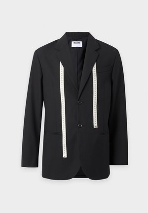 Man draagt een zwart colbert met meetlintdetail, bijpassende broek met ritszak, lichtblauw overhemd en zwarte loafers, staand tegen een witte achtergrond.