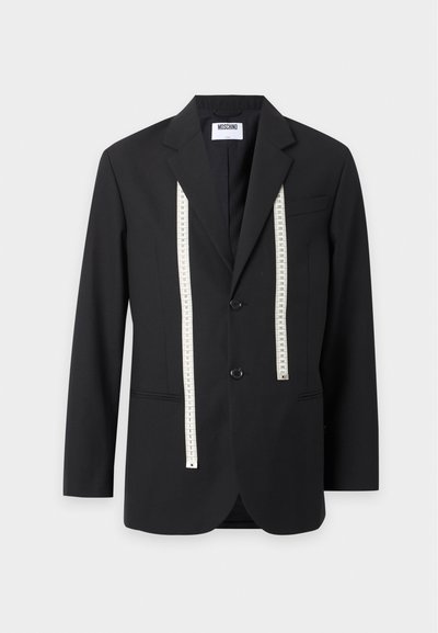 Veste noire Moschino avec deux boutons à l'avant et un ruban à mesurer blanc suspendu sur un fond clair uni.