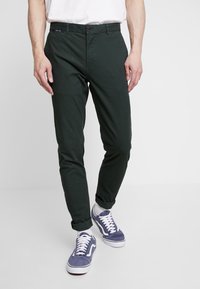 Zelené bavlněné kalhoty s úzkým střihem, zapínání na knoflíky a ohrnutými lemy. Kombinovány s modrými teniskami s bílými detaily.