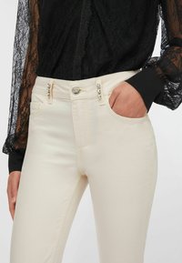 Beigenväriset kapeat farkut, joissa metallinen napin suljin ja koristeellisia kiviä etutaskuissa. Pehmeä, sileä kangaspinta.