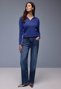 Femme debout avec une main dans la poche, portant un haut bleu à manches longues, un jean droit, des chaussures argentées pointues et un bracelet au poignet gauche.