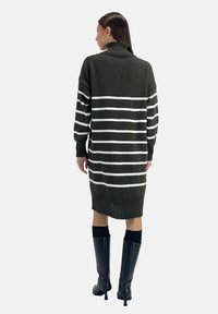 Olivgrünes gestricktes Pulloverkleid mit weißen horizontalen Streifen, Rollkragen, langen Ärmeln und geripptem Saum, kombiniert mit kniehohen schwarzen Stiefeln.