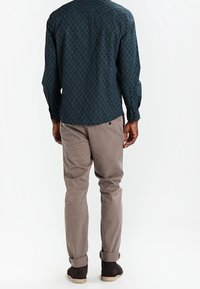 Langsleeve shirt in donker teal met geometrisch patroon, gecombineerd met lichtbeige broek. Schoenen zijn donkerbruin met een zachte textuur.