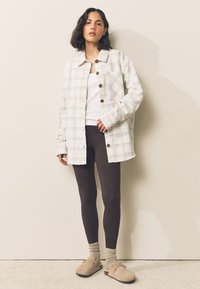 Kostkovaná košile na knoflíky světle béžové barvy s modrými a hnědými akcenty, doplněná hnědými legínami, vlněnými ponožkami a béžovými nazouvacími botami.