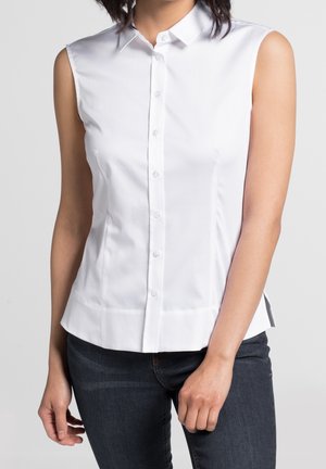 Vrouw draagt een mouwloos wit overhemd met knopen en donkerblauwe jeans, staand met een ontspannen houding tegen een lichte achtergrond.